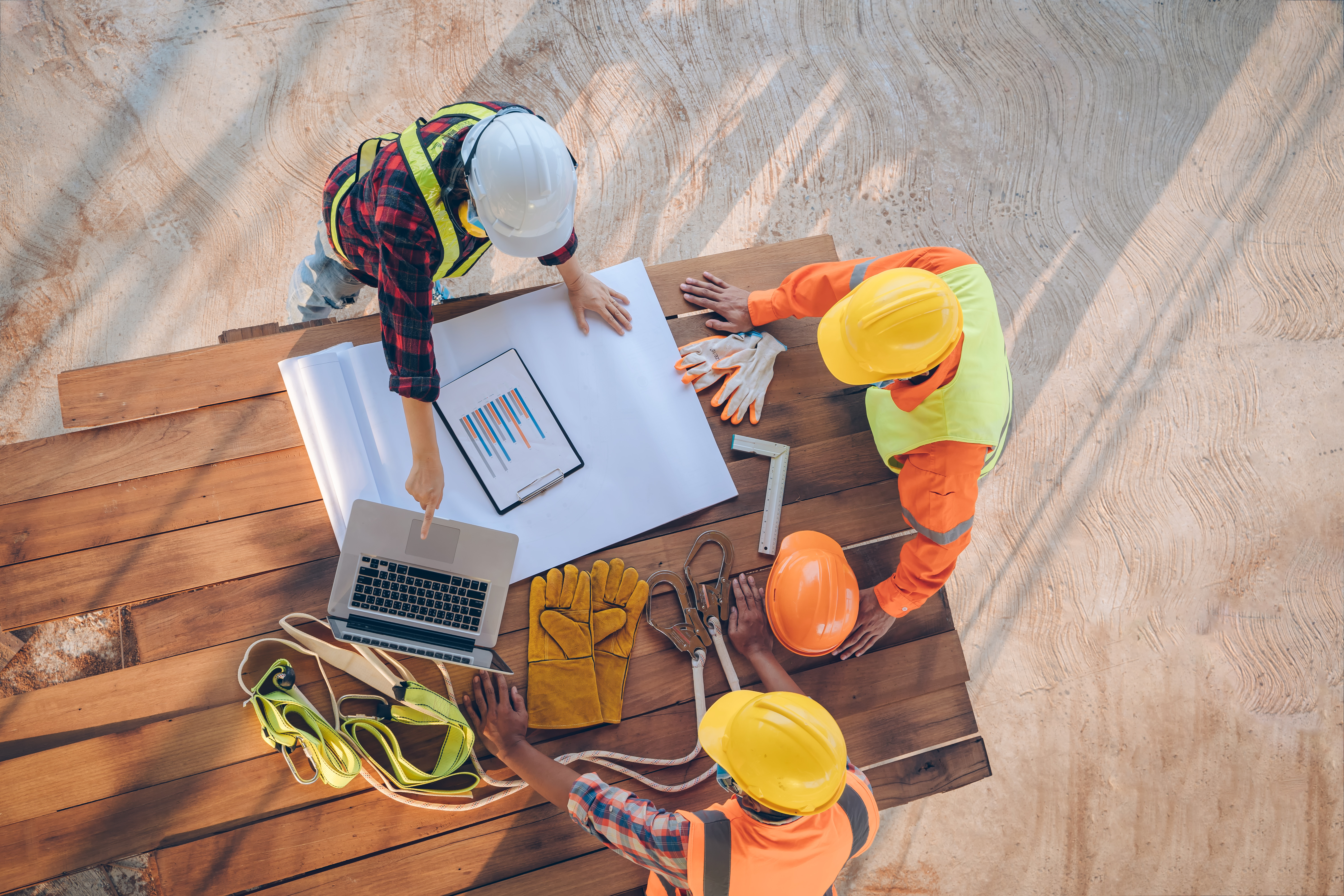 Three construction workers in hard hats and safety vests are seen reviewing blueprints at a construction site.