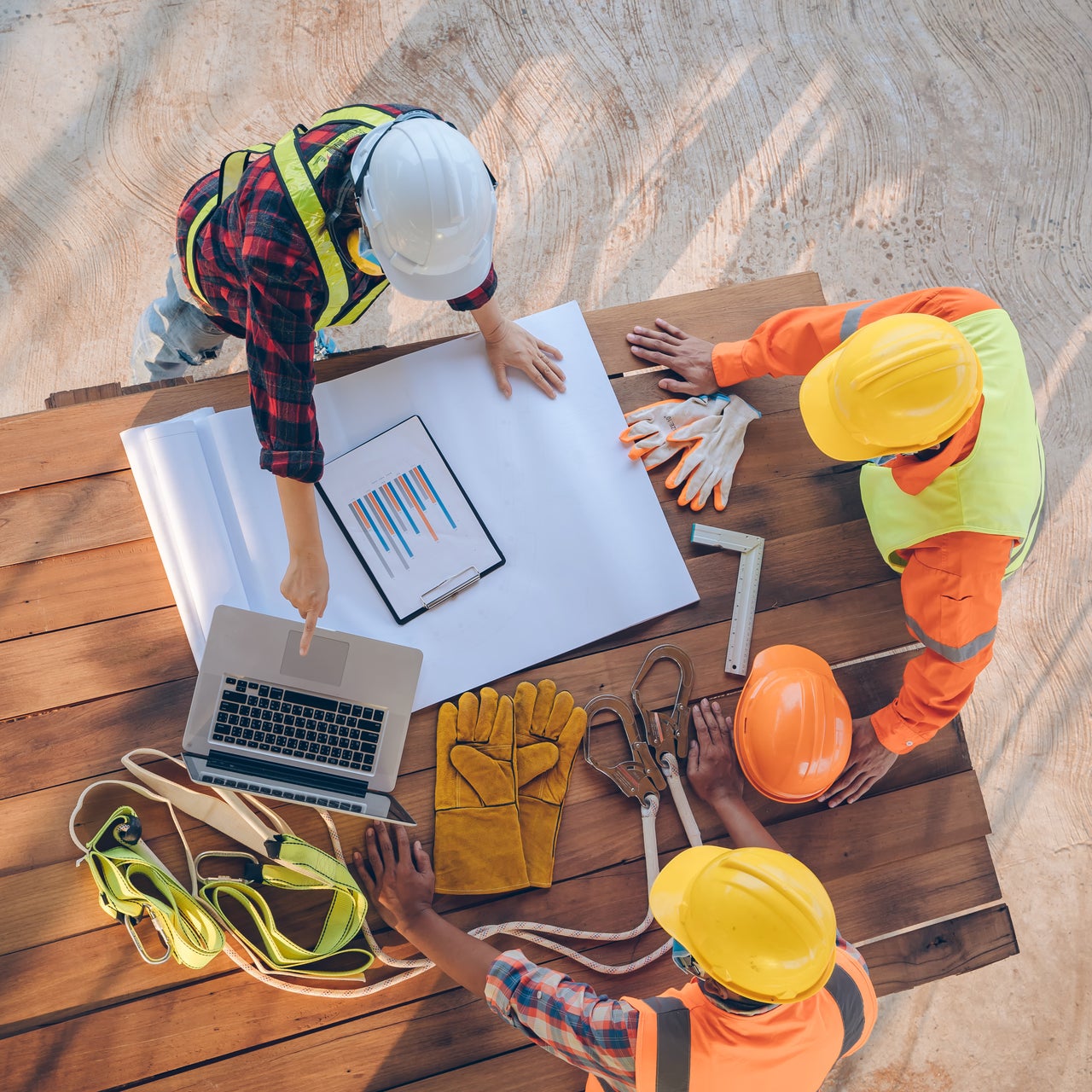 Three construction workers in hard hats and safety vests are seen reviewing blueprints at a construction site.