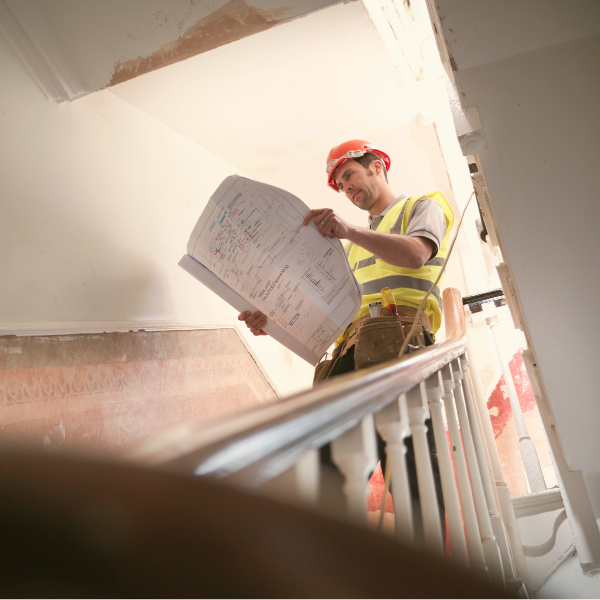 A construction worker stands on a staircase, reviewing building plans. 