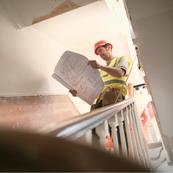 A construction worker stands on a staircase, reviewing building plans.