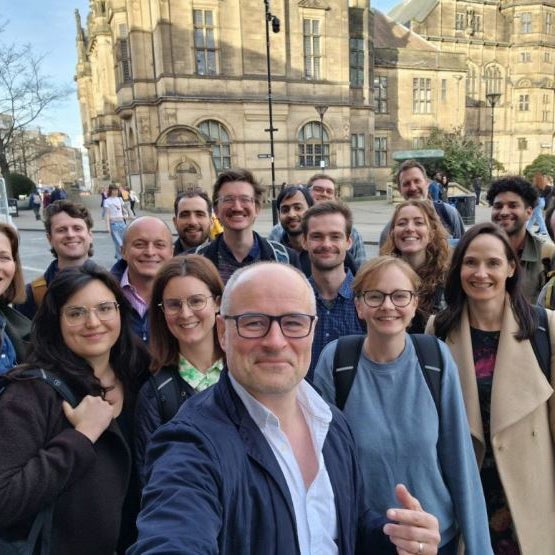A group of twelve people standing together on a stage or platform, posing for a group photo with raised arms and open gestures, celebrating or expressing excitement.