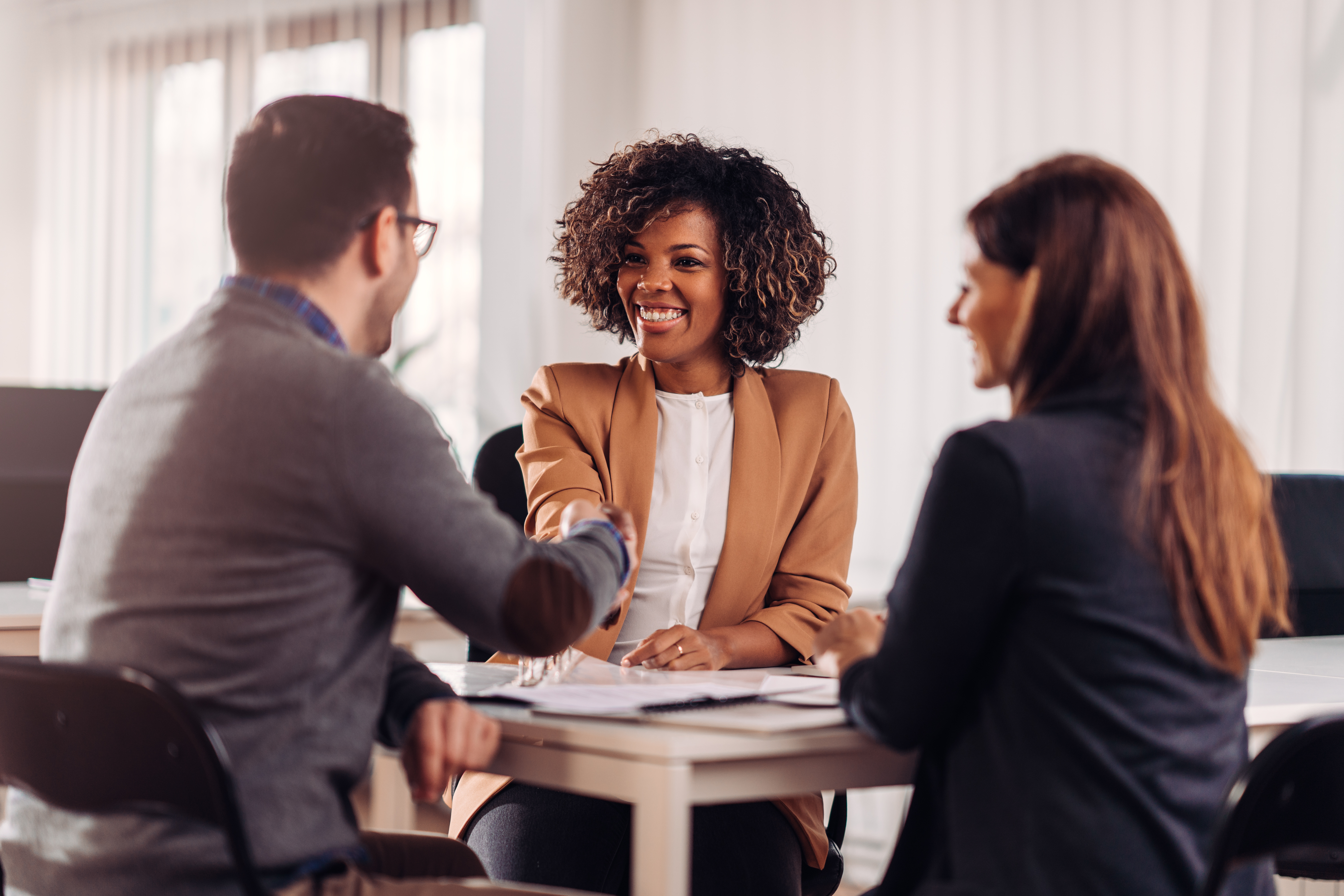 Two business people, a man and a woman, shaking hands across a table in an office setting.