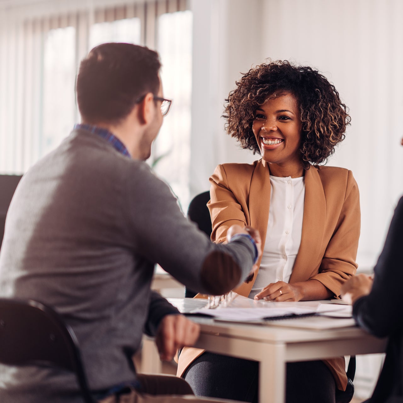 Two business people, a man and a woman, shaking hands across a table in an office setting.