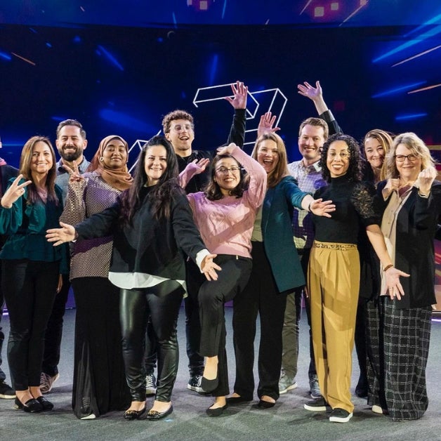 A group of twelve people standing together on a stage or platform, posing for a group photo with raised arms and open gestures, celebrating or expressing excitement.