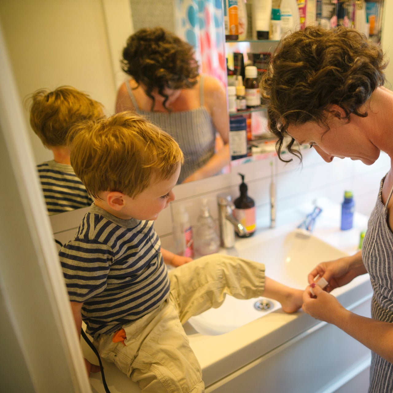 A mother helping her toddler put a bandage in one of his toes. The toddler is in a sitting position while the mother is standing.