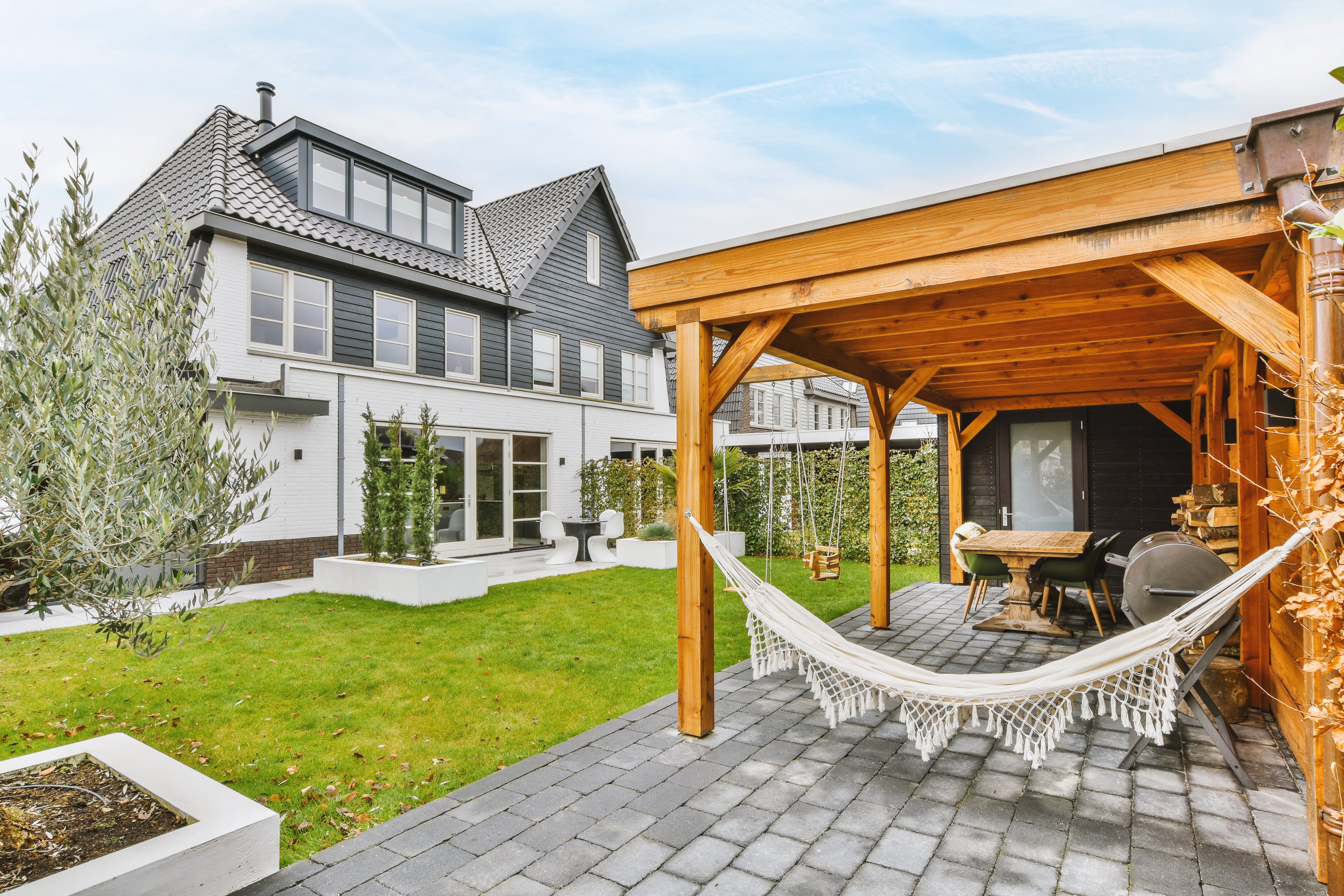 Paved patio with wooden roof and hammock located near grassy lawn against cloudy blue sky in backyard of suburban house.