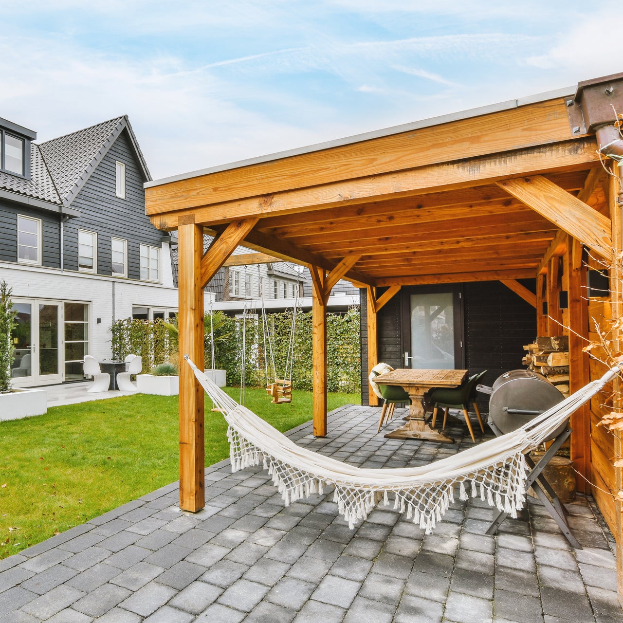 Paved patio with wooden roof and hammock located near grassy lawn against cloudy blue sky in backyard of suburban house.