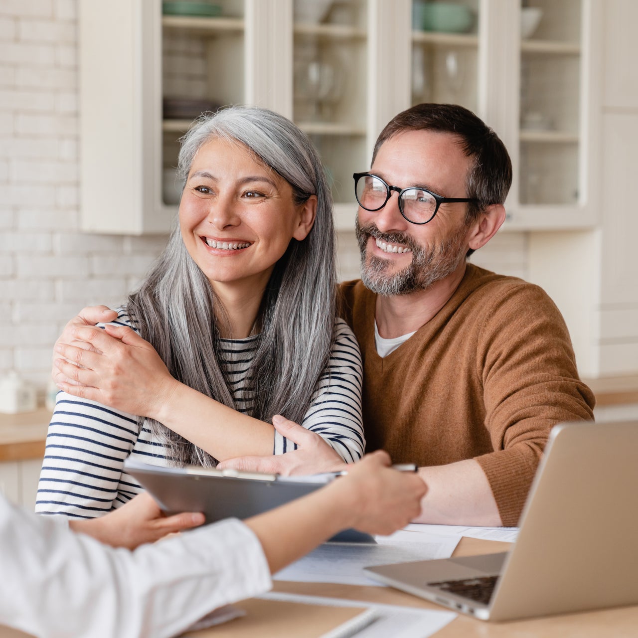An older couple is seated at a table, smiling and looking off to the side, while someone presents a document on a clipboard.