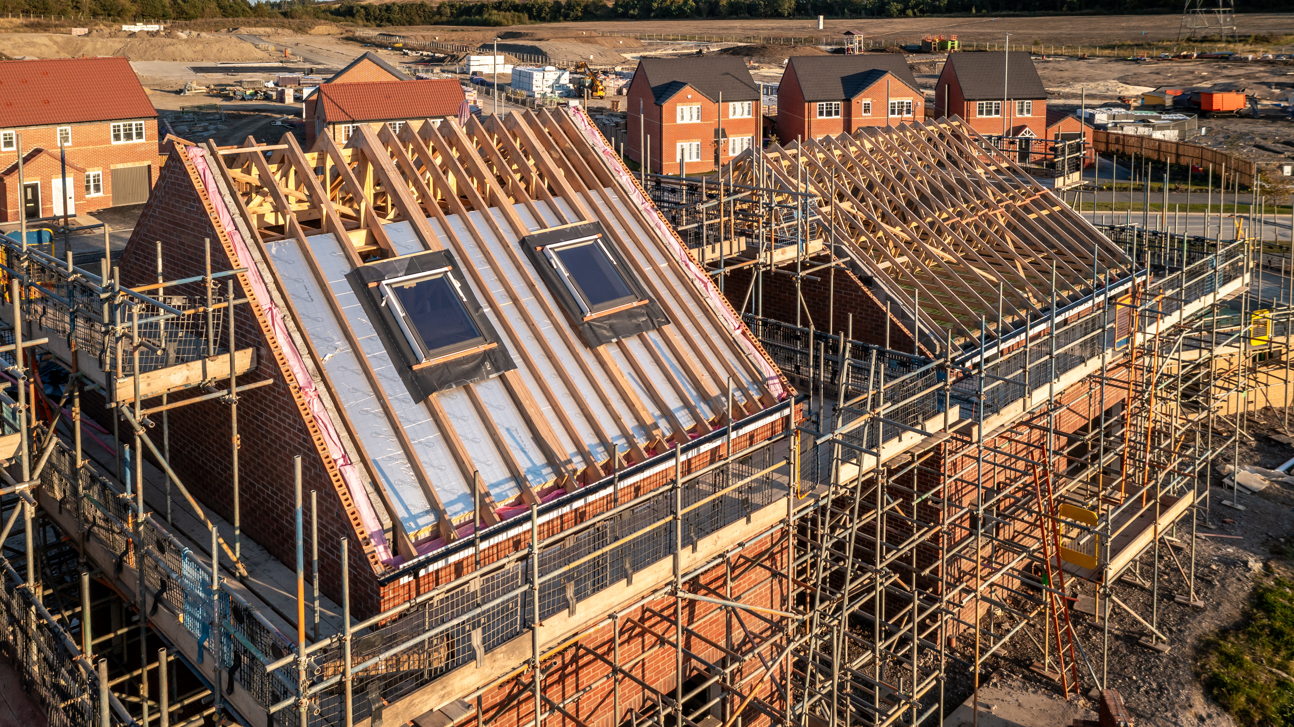 Aerial view of a roof under construction with beams and apex supporting exterior skylight windows on a new build house surrounded by scaffolding on a construction site.