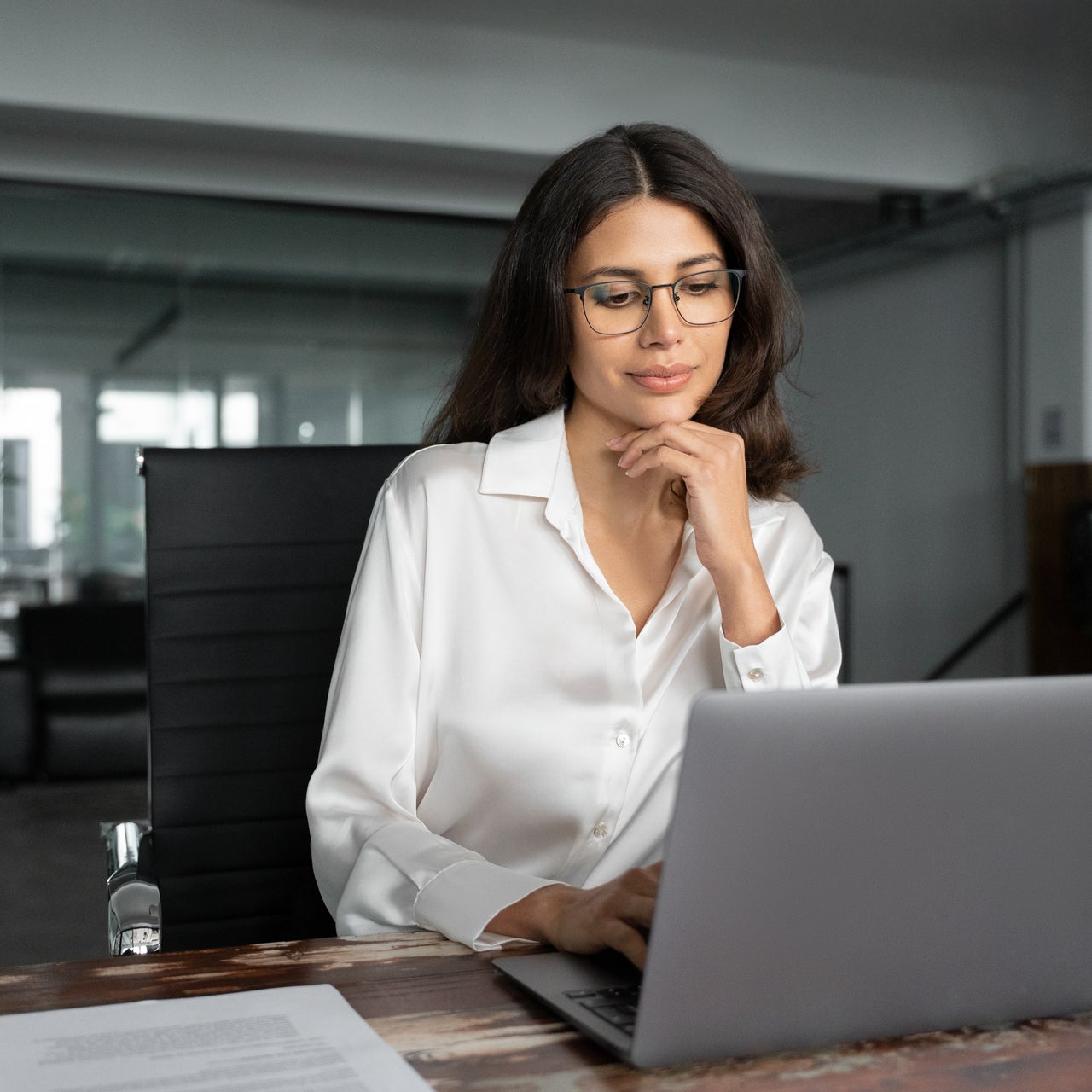 A focused young woman wearing glasses is sitting at a wooden desk, looking intently at a silver laptop with one hand resting on her chin.