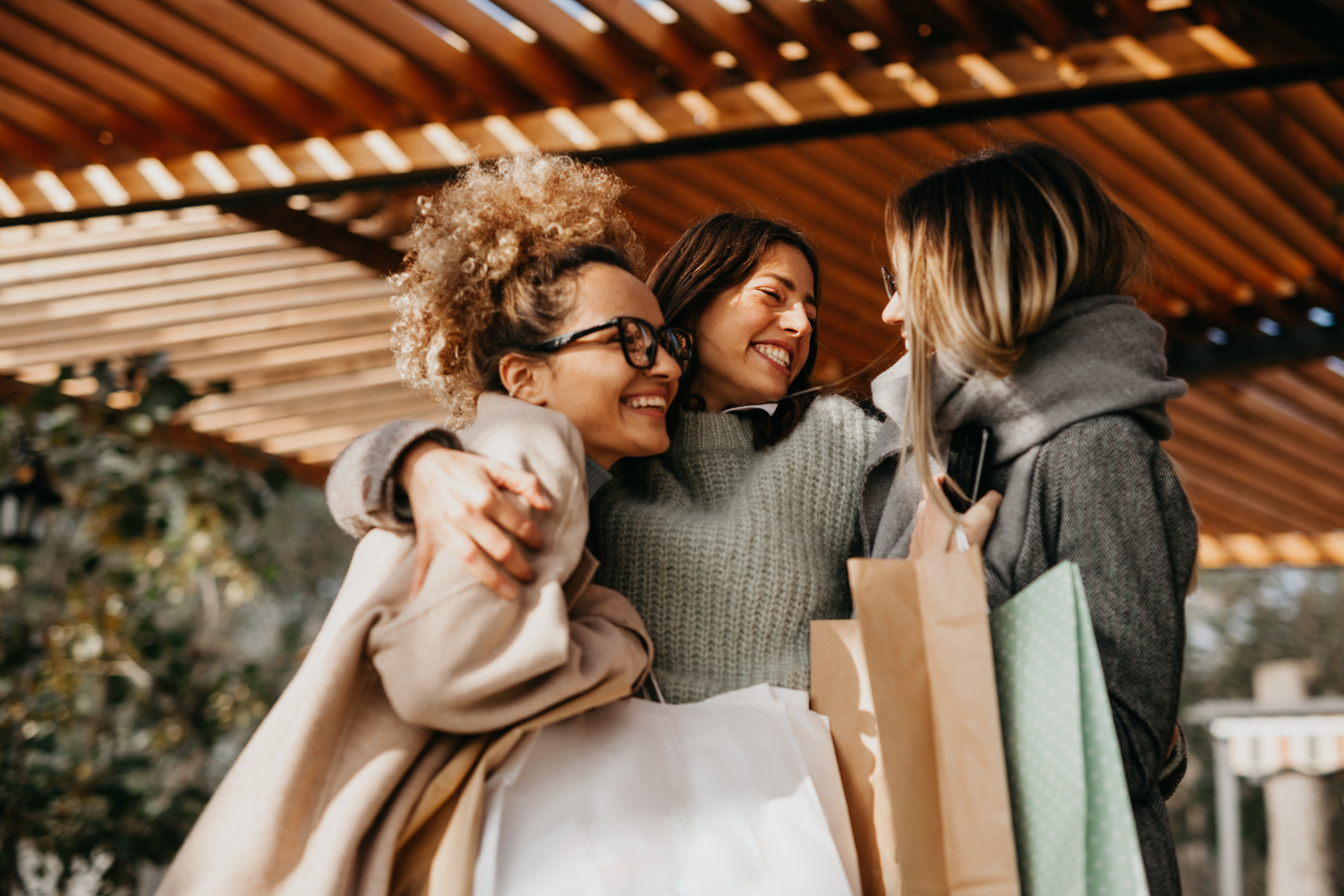 Three happy friends embracing with retail bags, celebrating a successful day of shopping.