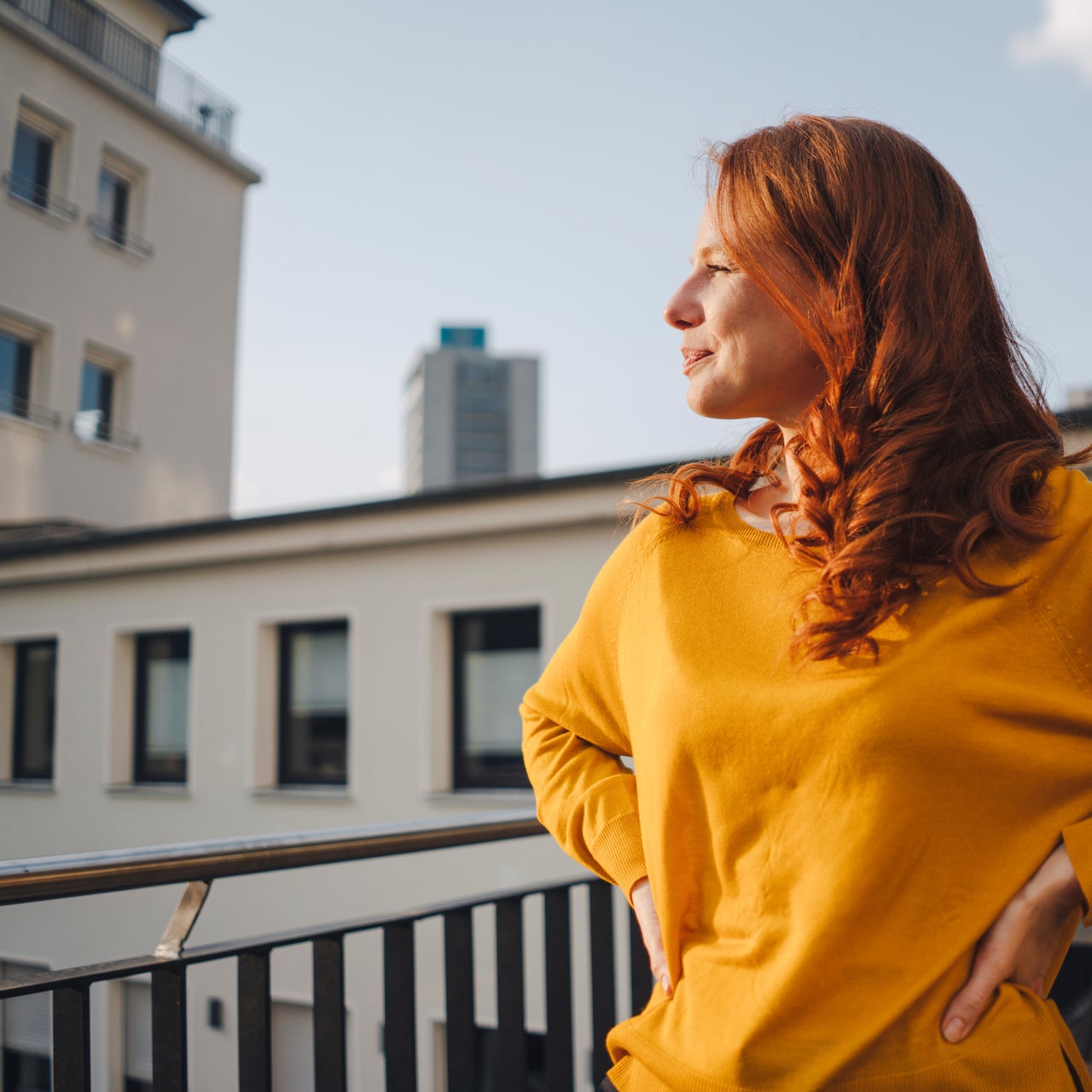 A woman with red hair, wearing a yellow sweater, is standing on a balcony.