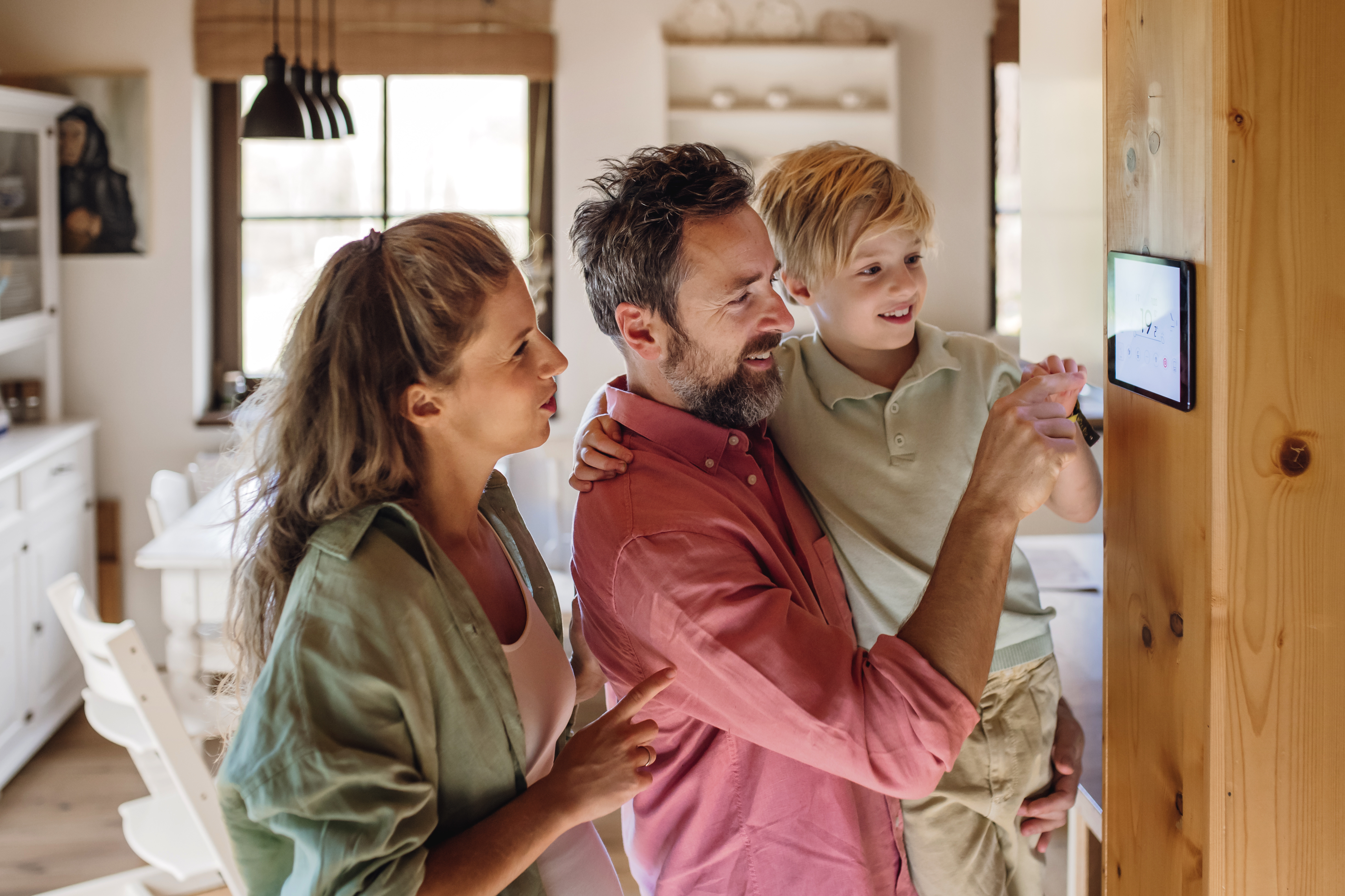 A family of three looks at a smart home display panel on a wall.