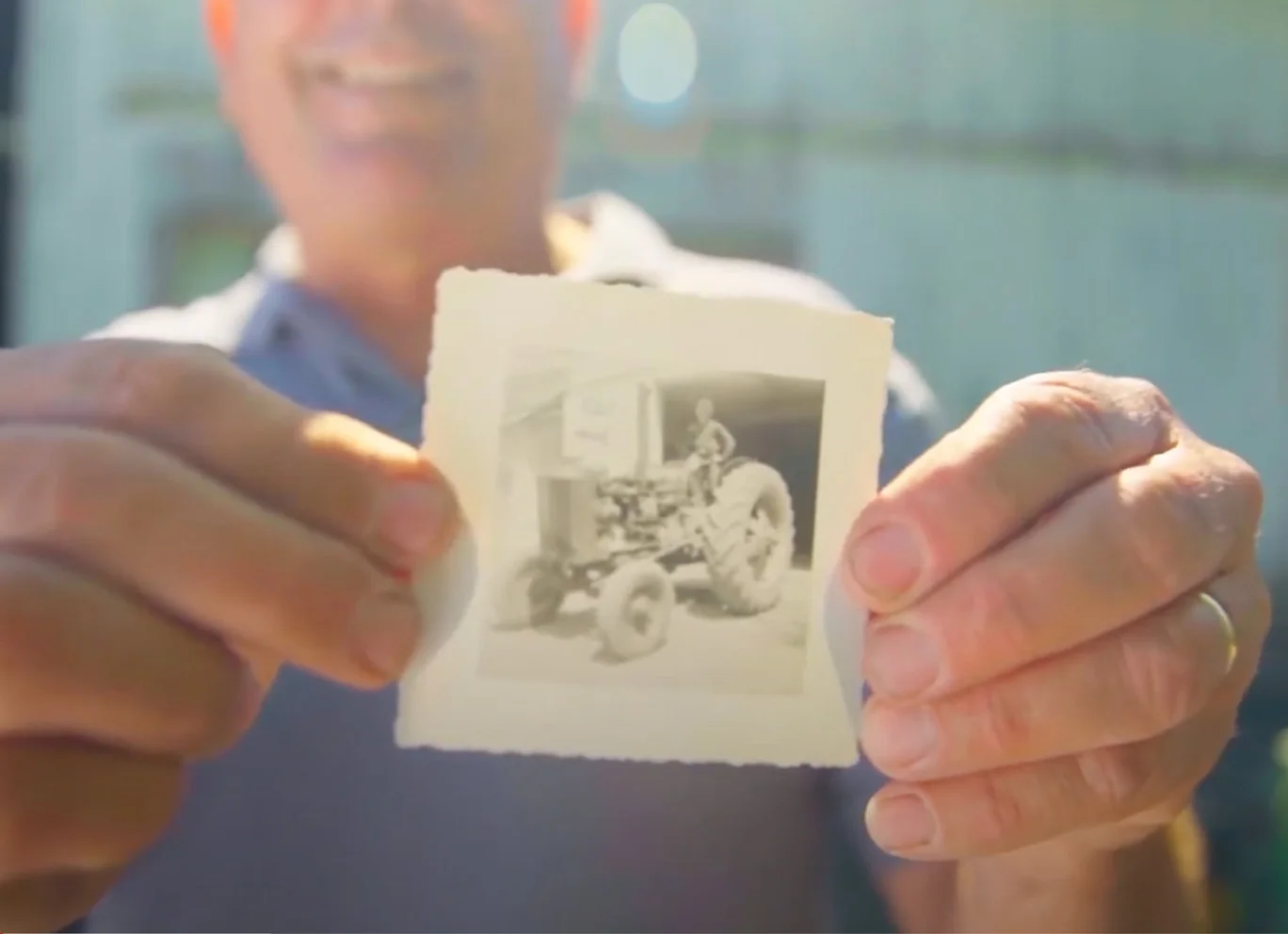A man holds an old photograph of a farmer on a tractor.