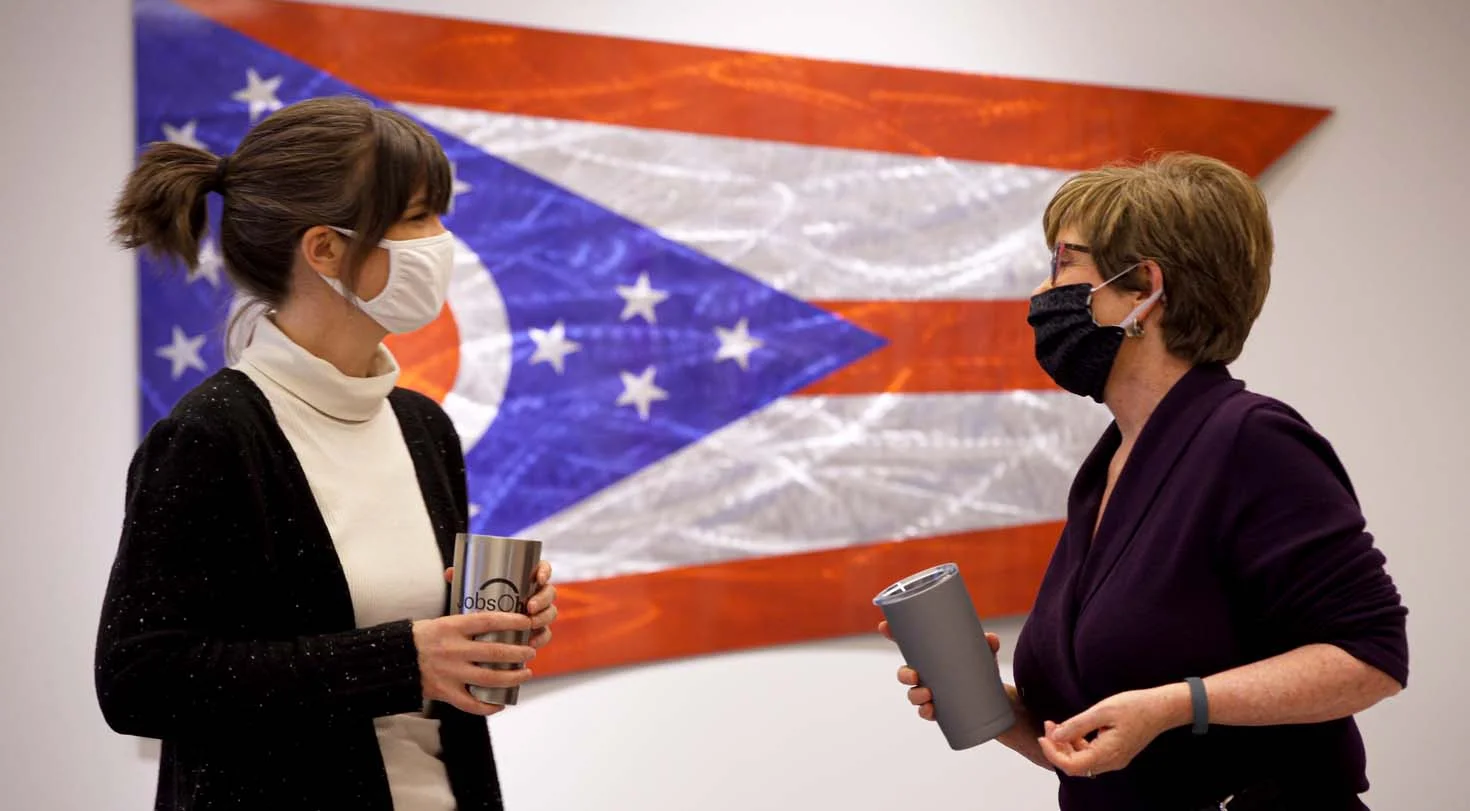 2 women wearing masks holding coffee cups with a flag in the background