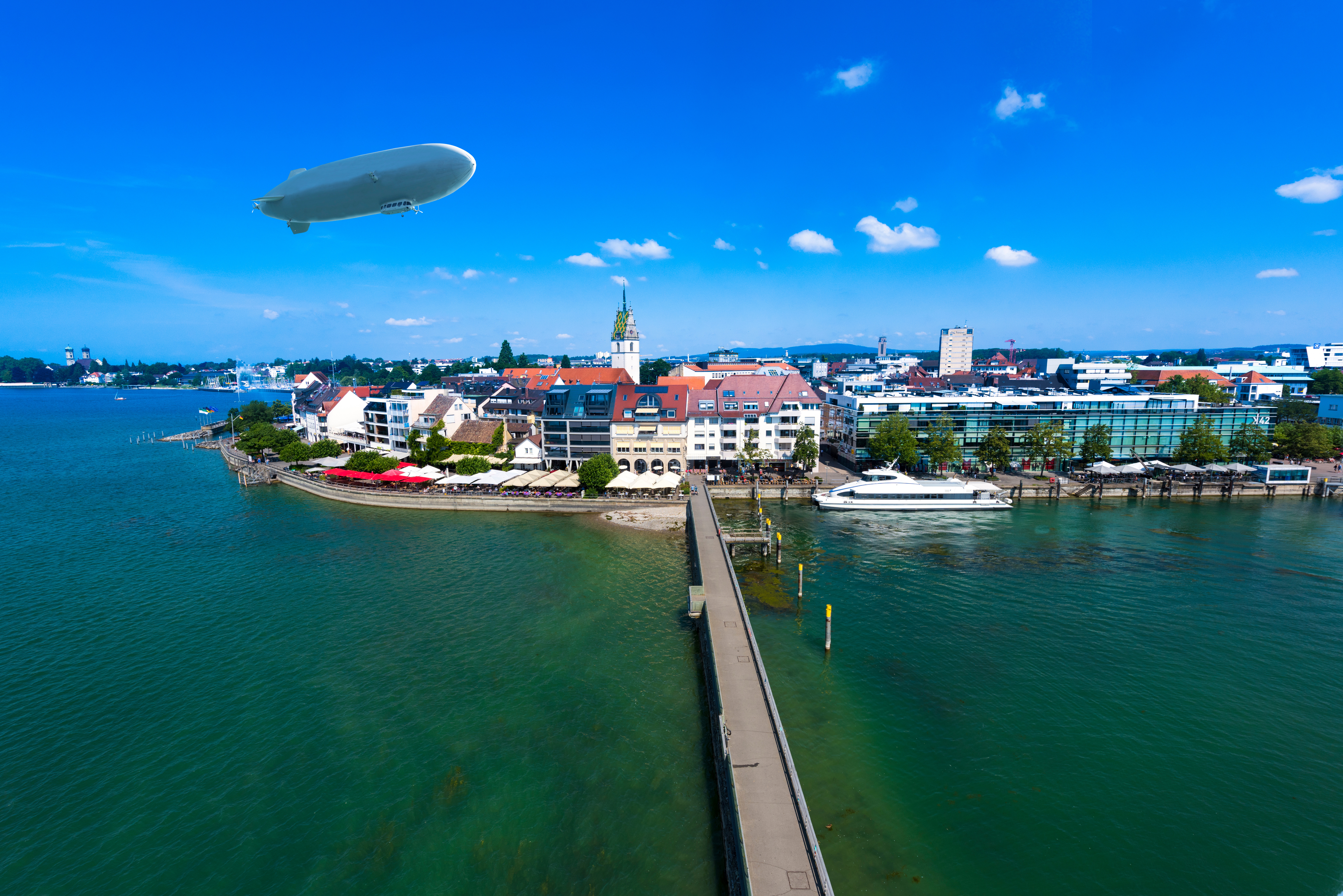 Aerial view of Friedrichshafen, Germany, featuring the Lake Constance waterfront, city buildings, a passenger ferry, and a Zeppelin in a blue sky. SHL Advantec will exhibit medical injection molds and autoinjector tooling expertise at Fakuma 2026.