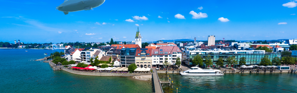 Aerial view of Friedrichshafen, Germany, featuring the Lake Constance waterfront, city buildings, a passenger ferry, and a Zeppelin in a blue sky. SHL Advantec will exhibit medical injection molds and autoinjector tooling expertise at Fakuma 2026.