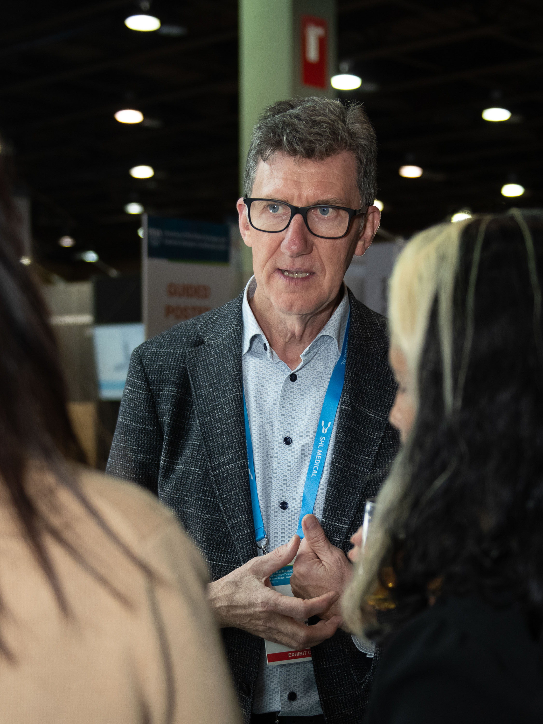 Man wearing glasses and a blazer speaking with two visitors at an event. SHL Medical’s Head of Business Development Europe, the Middle East, and Africa highlights cost-effectiveness as central to European self-injection markets.