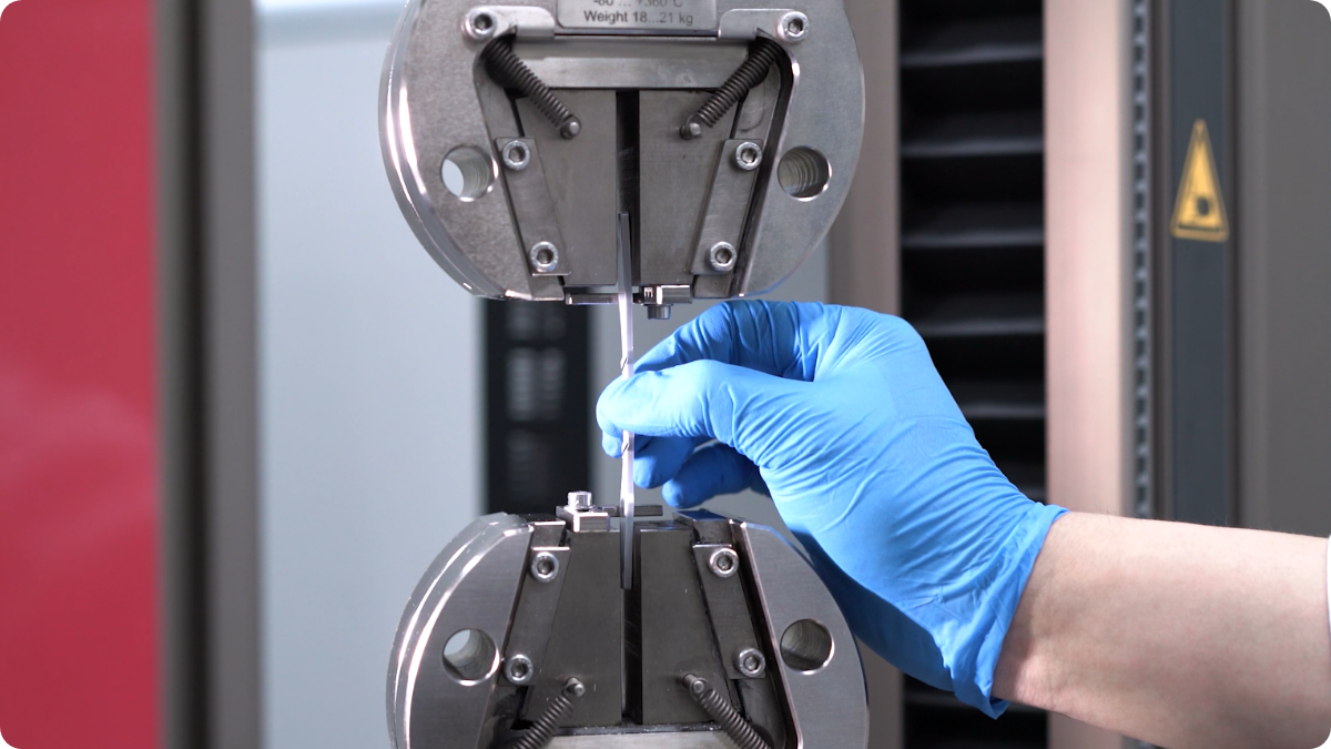 A gloved hand places a small test specimen into a tensile testing machine.