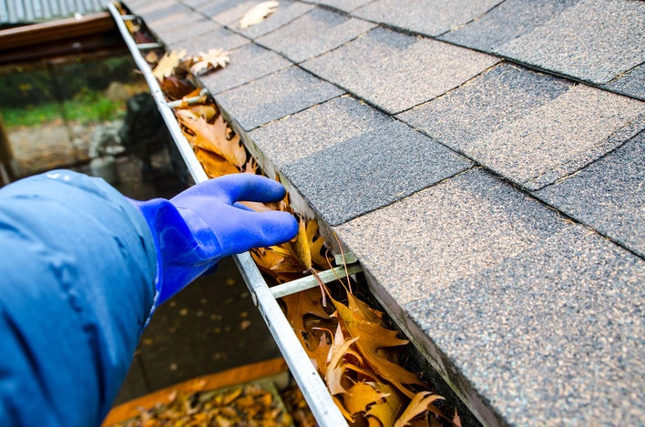 A gloved hand cleaning leaves and debris from gutters for home winterization.