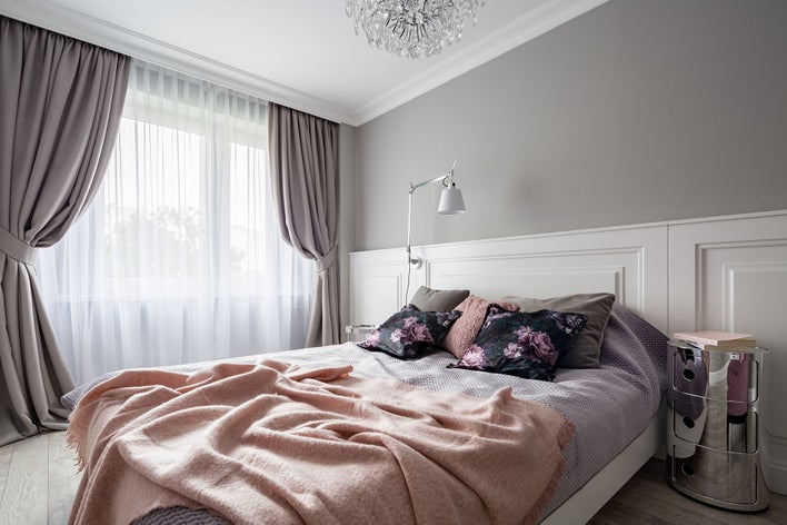 A cozy bedroom with purple and gray tones throughout that has floor-to-ceiling curtains covering the large windows.