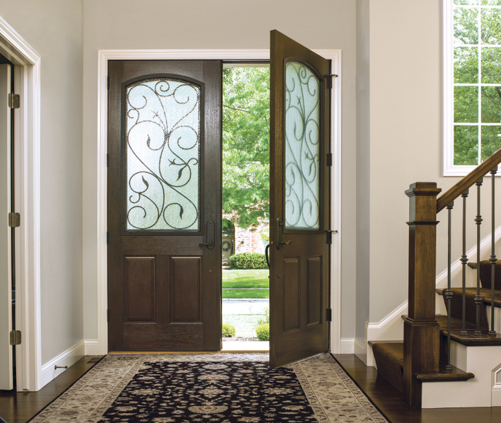 An interior view of a pair of woodgrain fiberglass doors with decorative privacy glass<br>panels opening to a sunny exterior at a home’s entrance.