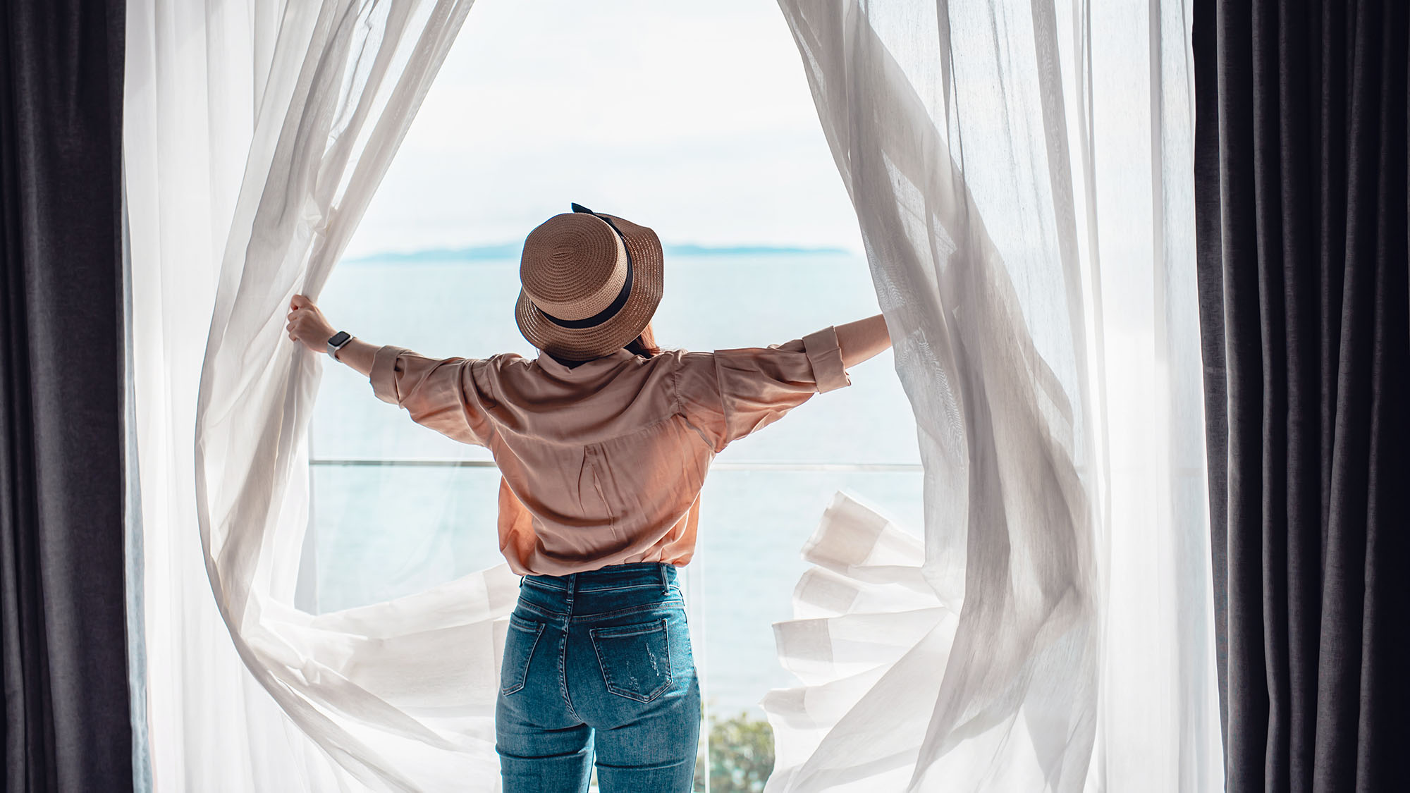 A woman increasing the natural light in a room by opening curtains. There is an ocean view beyond.