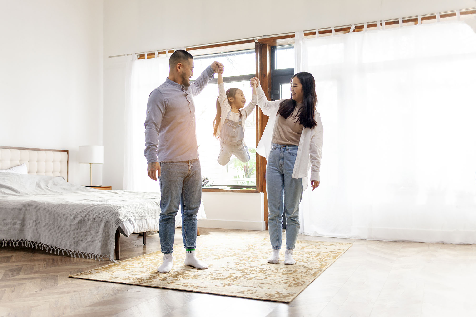 Parents lifting their child up by the arms in a sunlit bedroom.