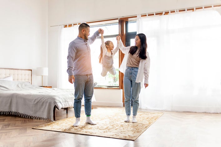 Parents lifting their child up by the arms in a sunlit bedroom.
