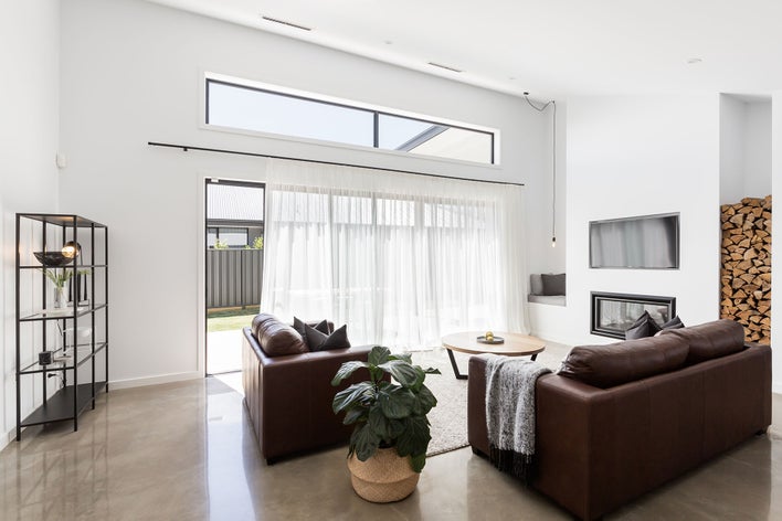 A modern living room with tile flooring and brown furniture that is naturally lit from a wall of large sliding glass patio doors that are covered with sheer curtains.