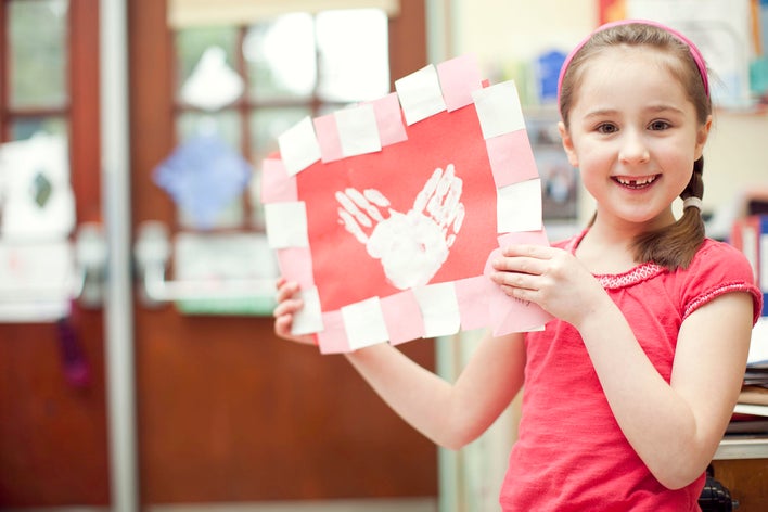 A young girl showcasing her Valentine’s Day decoration with handprints in white paint onred construction paper, mimicking the shape of a heart.