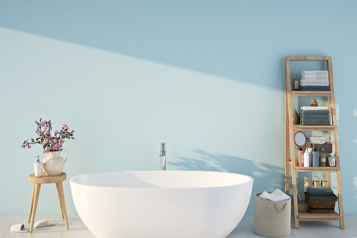 A free-standing white bathtub and wood bathroom furniture against a light blue wall