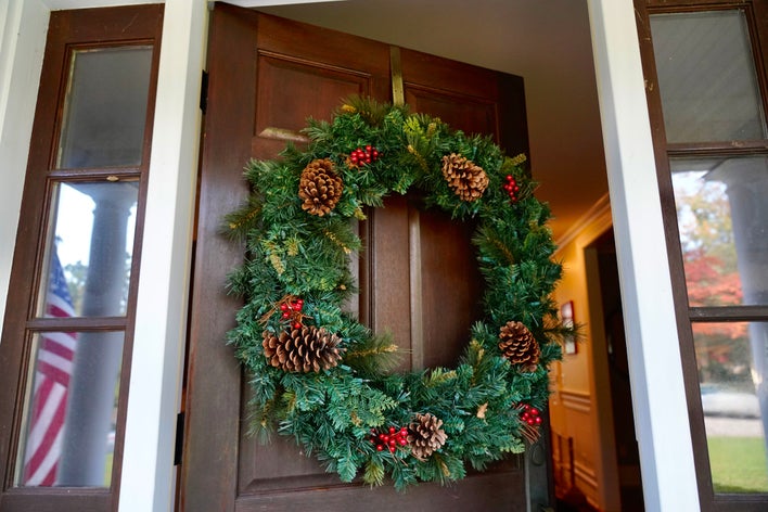 A pinecone and red berry decorated wreath hanging on an open woodgrain entry door with a brass over-the-door wreath hanger.