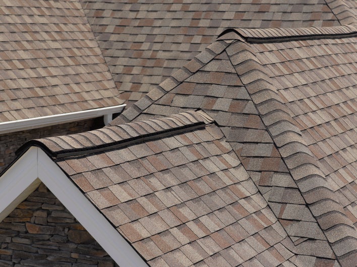 Multi-colored shingles on the gabled roof of a house with stacked stone exterior and white gutters.