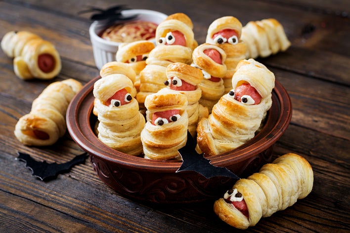 : A Halloween-themed dish of mummy-themed pigs in a blanket inside a haunted house