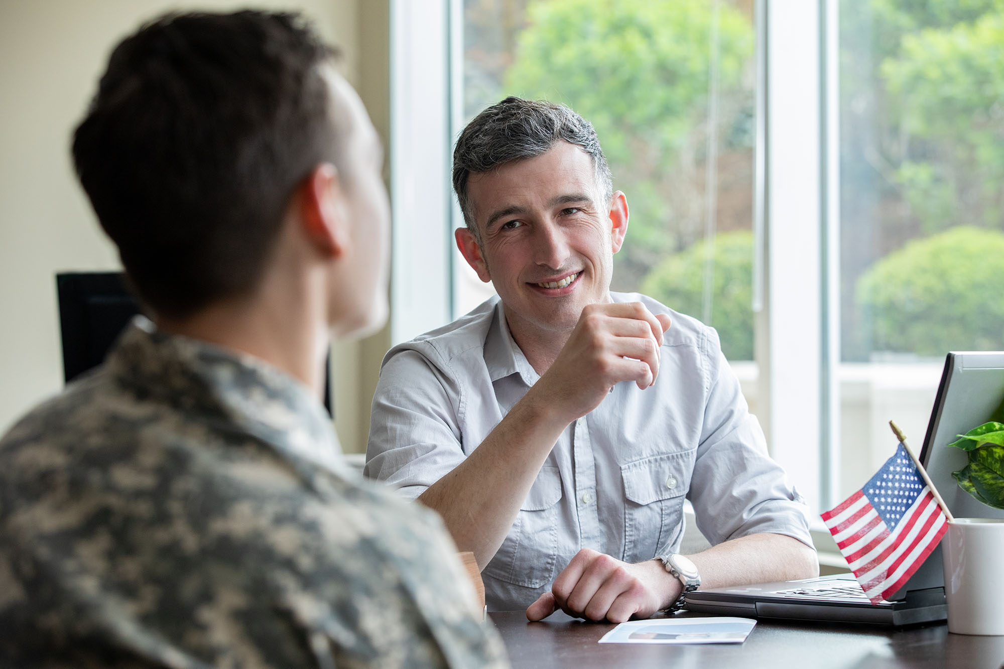 Two men talking together at a table with a small American flag present. One man is in a military uniform, and the other is in civilian clothes. 