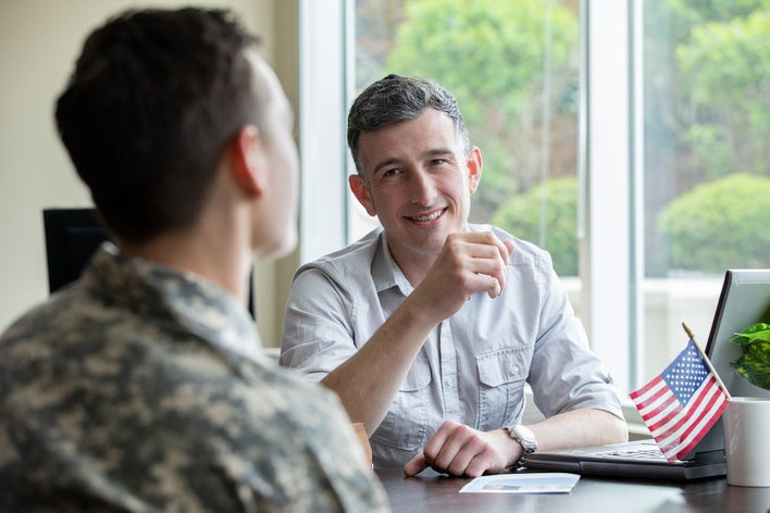 Two men talking together at a table with a small American flag present. One man is in a military uniform, and the other is in civilian clothes.