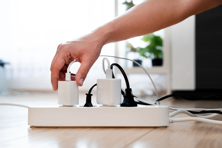 A surge protector with several cords and chargers plugged into it. A hand is reaching in to unplug a device not currently in use.