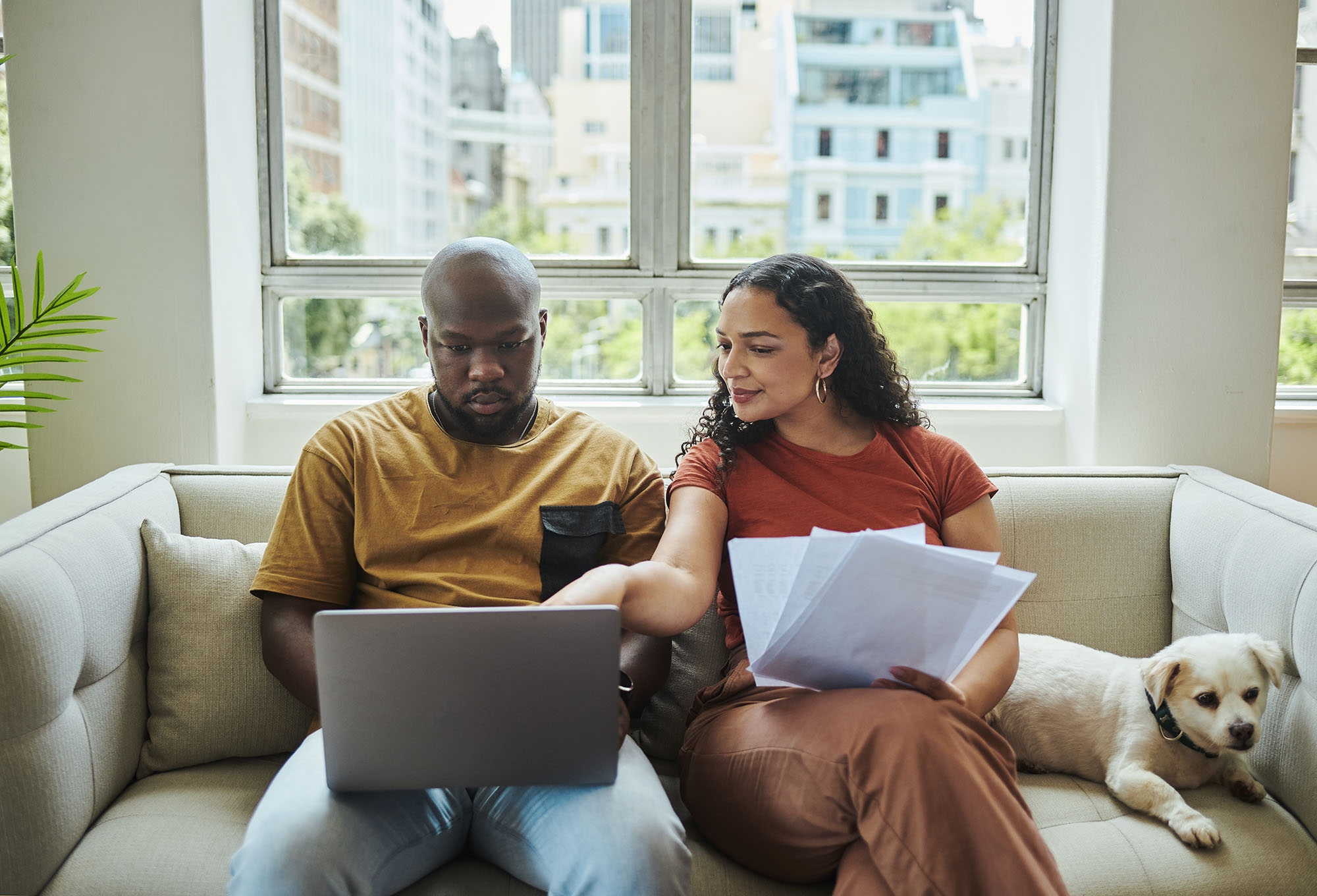 Man and woman sitting on a couch with a dog as they review their new home checklist on a laptop.