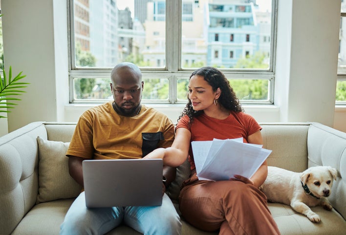 Man and woman sitting on a couch with a dog as they review their new home checklist on a laptop.