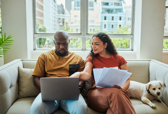 Man and woman sitting on a couch with a dog as they review their new home checklist on a laptop.