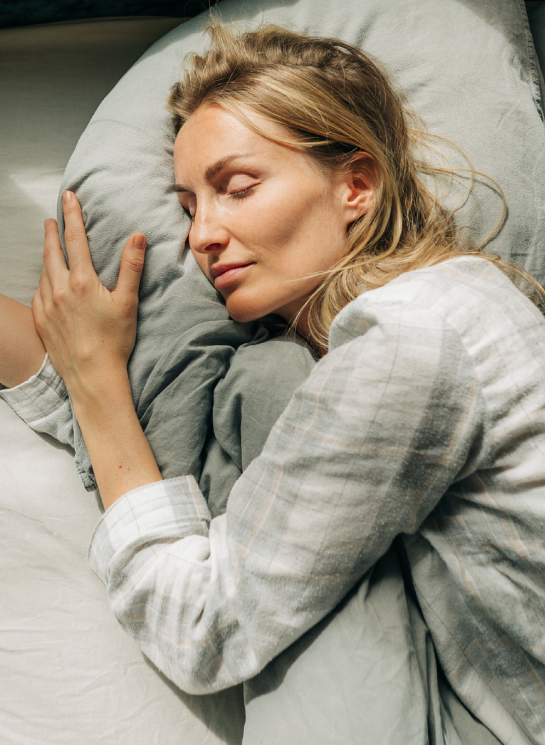 A woman with blonde hair rests peacefully on her side, hugging a pillow tightly while lying on a bed with grey sheets.
