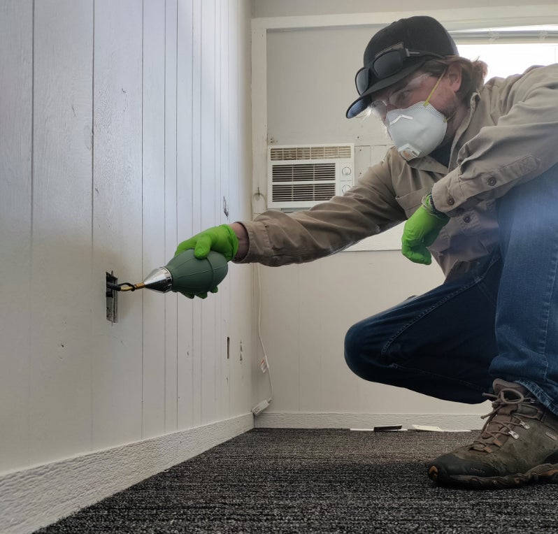 Pest control professional treating wall cracks with protective gear and equipment.