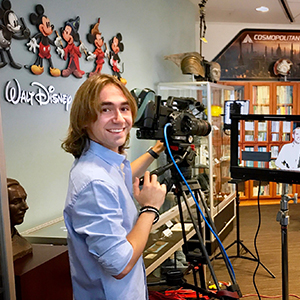 Male Disney Intern with blonde hair and a blue collared shirt smiling while standing behind a film camera