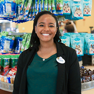 Female Disney Intern with shoulder length black hair, a green shirt and black jacket smiling in front of a store display of Disney snacks