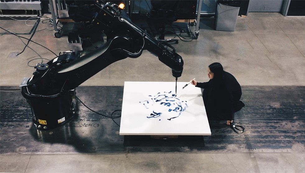 Woman collaborating with large robotic arm to paint on canvas in an industrial studio setting.