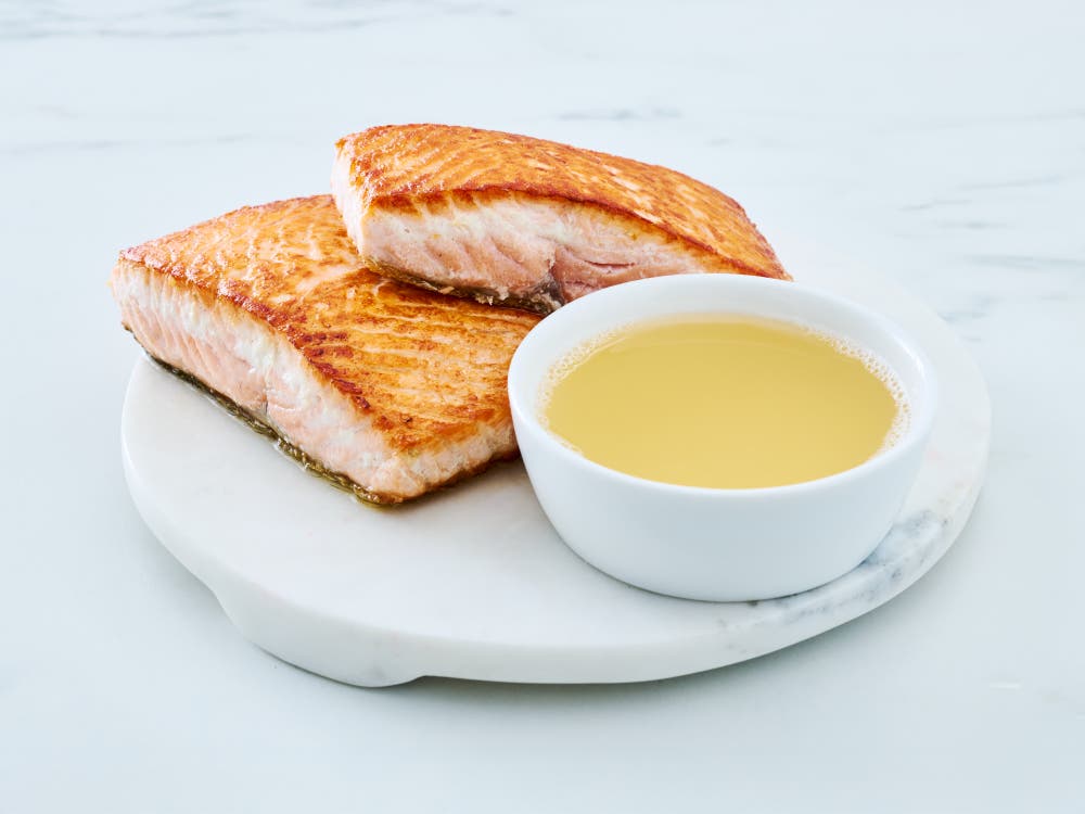 Wild Alaska salmon broth meal supplement in a white bowl, styled on a round white marble board beside two cooked salmon fillets, showing a clear, golden broth on a light background.