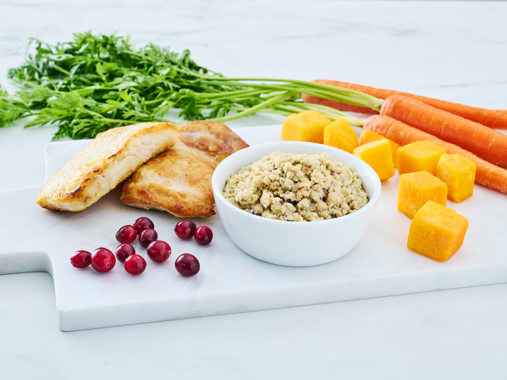 Wild Alaska pollock and chicken minced cat food in a white bowl, styled on a white cutting board with cooked chicken and fish fillets, whole carrots with greens, cubed squash, and fresh cranberries.