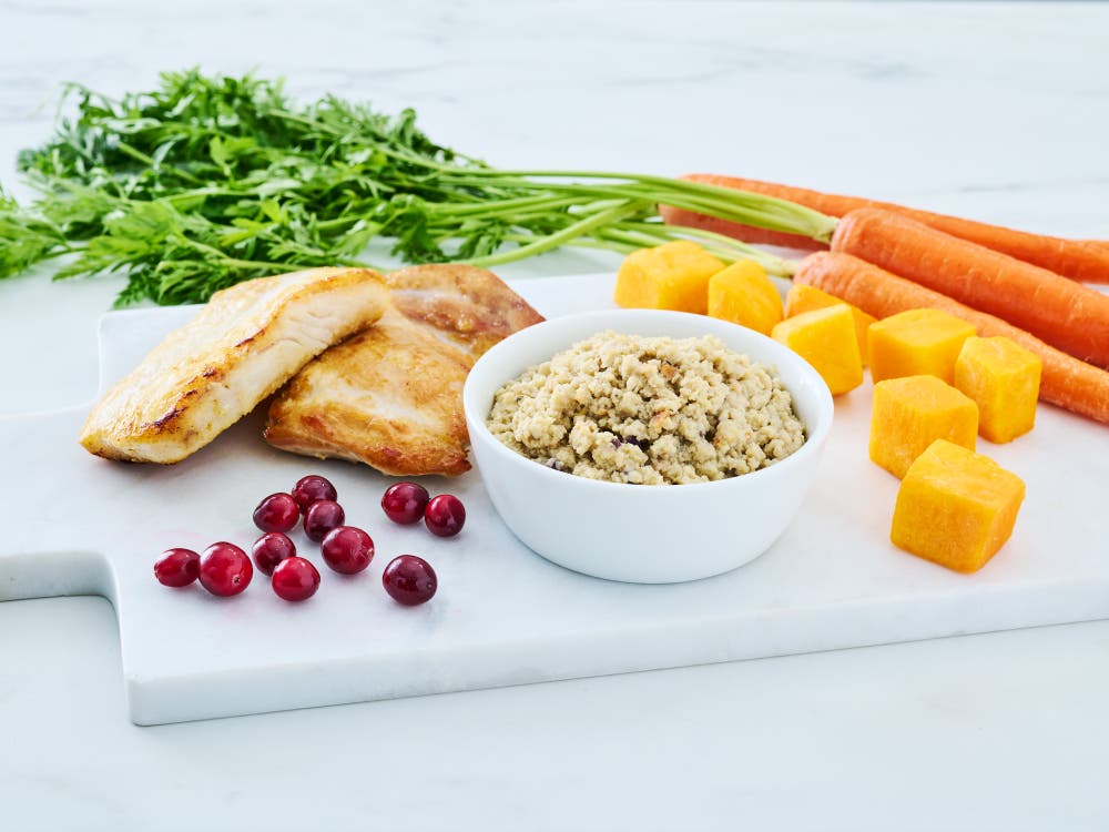 Wild Alaska pollock and chicken minced cat food in a white bowl, styled on a white cutting board with cooked chicken and fish fillets, whole carrots with greens, cubed squash, and fresh cranberries.