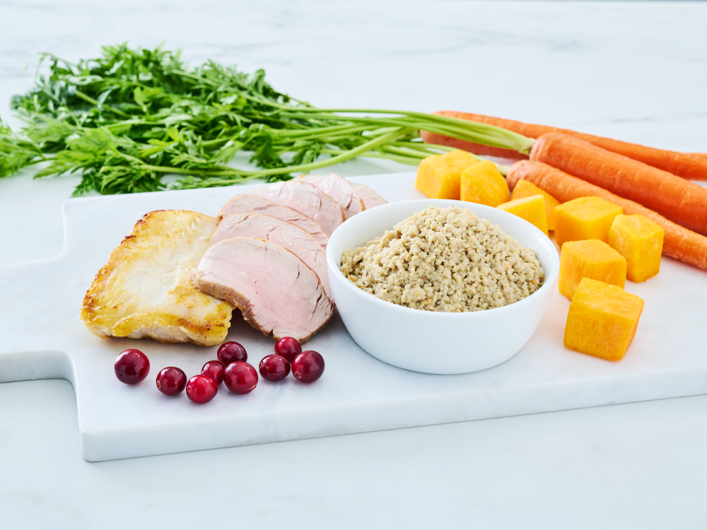 Wild Alaska pollock and pork minced cat food in a white bowl, styled on a white cutting board with sliced pork, a cooked fish fillet, carrots, leafy greens, cubed squash, and cranberries.